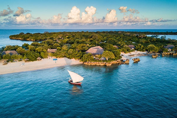 Aerial view of a remote island in Mozambique with pristine beaches, lush greenery, and a traditional dhow sailboat, offering the ultimate luxury safari experience.