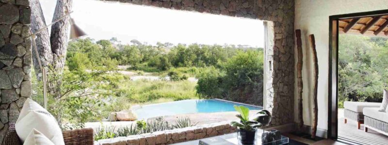 Interior view of a luxurious safari suite with a large open window looking out onto a private plunge pool and the natural savanna landscape beyond. The room features stone walls, comfortable seating, and a coffee table, providing a seamless connection with the outdoors.
