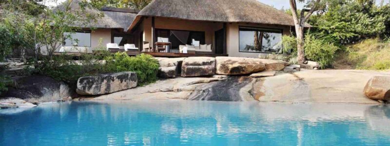 A luxurious safari lodge suite with a thatched roof, situated on the banks of a blue body of water, likely the Sand River. The suite blends into the natural environment with trees and sandy banks around it, suggesting a private and serene setting.