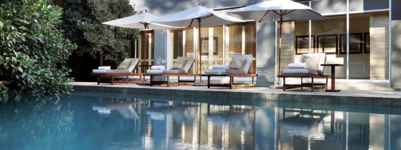 A serene outdoor swimming pool reflecting the elegant facade of the Saxon Hotel & Spa. Lounge chairs with white umbrellas are lined up along the poolside, surrounded by lush greenery, suggesting a luxurious and relaxing environment.