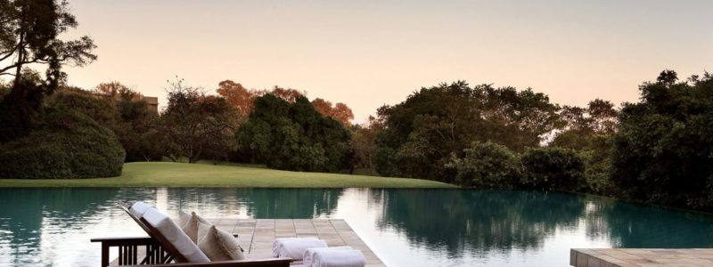 A serene outdoor pool area at the Saxon Hotel & Spa at dusk, featuring comfortable lounge chairs on a paved deck beside a reflective pool that appears to blend into a natural pond or water feature, surrounded by lush trees and a soft sunset sky.
