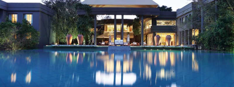 The grand exterior of the Saxon Hotel & Spa at dusk, featuring a large, illuminated building with a distinctive dome structure, reflected perfectly in a calm, expansive swimming pool in the foreground. Lush trees surround the elegant property.