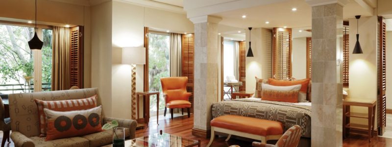 A luxurious and spacious hotel suite at the Saxon Hotel & Spa, featuring a king-sized bed, a comfortable sitting area with a sofa and armchairs, and elegant furnishings. The room is well-lit with natural light from large windows and tasteful lighting fixtures, showcasing a sophisticated design.