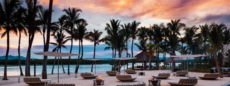 A stunning sunset view over the One&Only Le Saint Géran resort, showcasing a poolside lounge area with numerous elegant daybeds and umbrellas, silhouetted palm trees, and the tranquil lagoon under a vibrant, colorful sky.