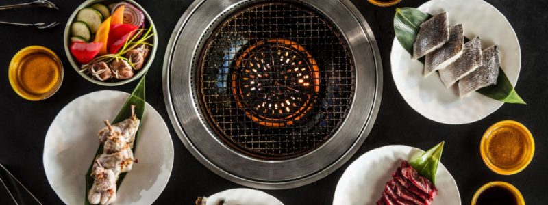 An overhead shot of a Korean BBQ setup, featuring a central grill surrounded by various plates of raw meats (like sliced beef and pork belly), fresh vegetables, sauces, and small dishes, suggesting a vibrant and interactive dining experience.