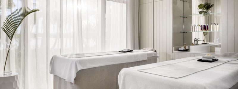 A serene and brightly lit couples' treatment room at the One&Only Le Saint Géran spa, featuring two massage beds draped in white linens. The room has large windows with sheer curtains and minimalist decor, creating a relaxing ambiance for wellness treatments.