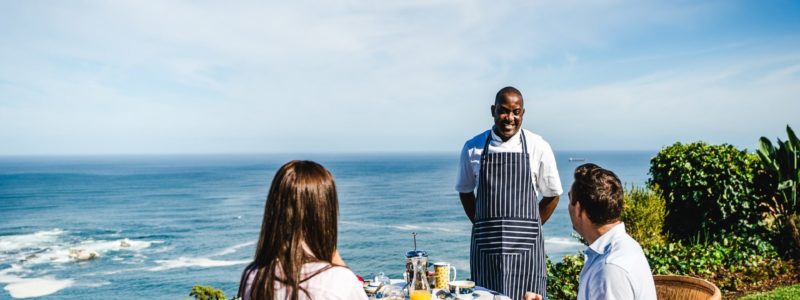 Couple enjoying a gourmet outdoor lunch with a chef at Cape Point Peninsula, overlooking the ocean on a sunny day.