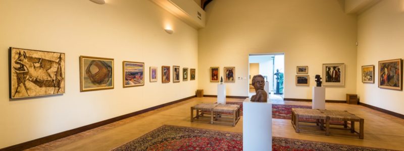 An interior view of an art gallery featuring numerous framed paintings on light-colored walls and several sculptures on pedestals. A large ornate rug covers the wooden floor in the center of the room, and the ceiling has exposed wooden beams.