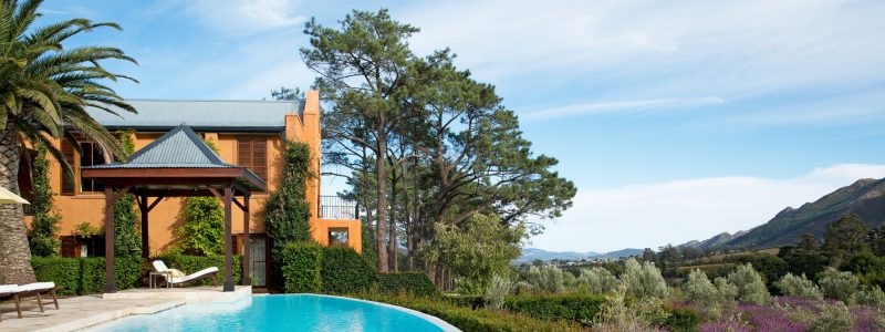 A luxurious resort in Africa with a curved swimming pool in the foreground. A building with an orange facade and a covered entrance is visible behind the pool, surrounded by lush green foliage and palm trees. In the background, there's a scenic view of rolling hills under a partly cloudy blue sky.