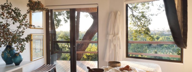 An inviting interior space at Londolozi's Healing House, set up for a spa or wellness experience. It features a massage bed with neatly folded towels, a singing bowl, and large windows and doors that offer a view of the natural surroundings.