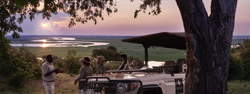 One guide and two tourists standing beside a safari jeep under a tree at sunset, overlooking a scenic river landscape, in the Chobe National Park, in Botswana.