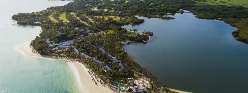 An aerial view of the One&Only Le Saint Géran resort situated on a lush green private peninsula in Mauritius, surrounded by the azure Indian Ocean on one side and a tranquil lagoon on the other. The resort's buildings, golf course, and white sandy beaches are visible, showcasing its idyllic location.