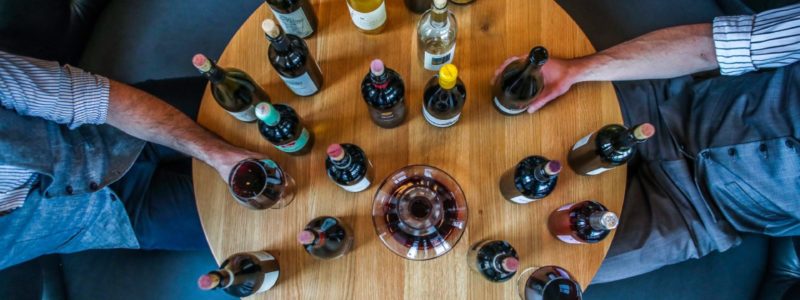 An aerial view of a wooden round table with several wine bottles and glasses, suggesting a wine tasting experience. Two people's arms are visible on either side of the table, holding wine glasses.
