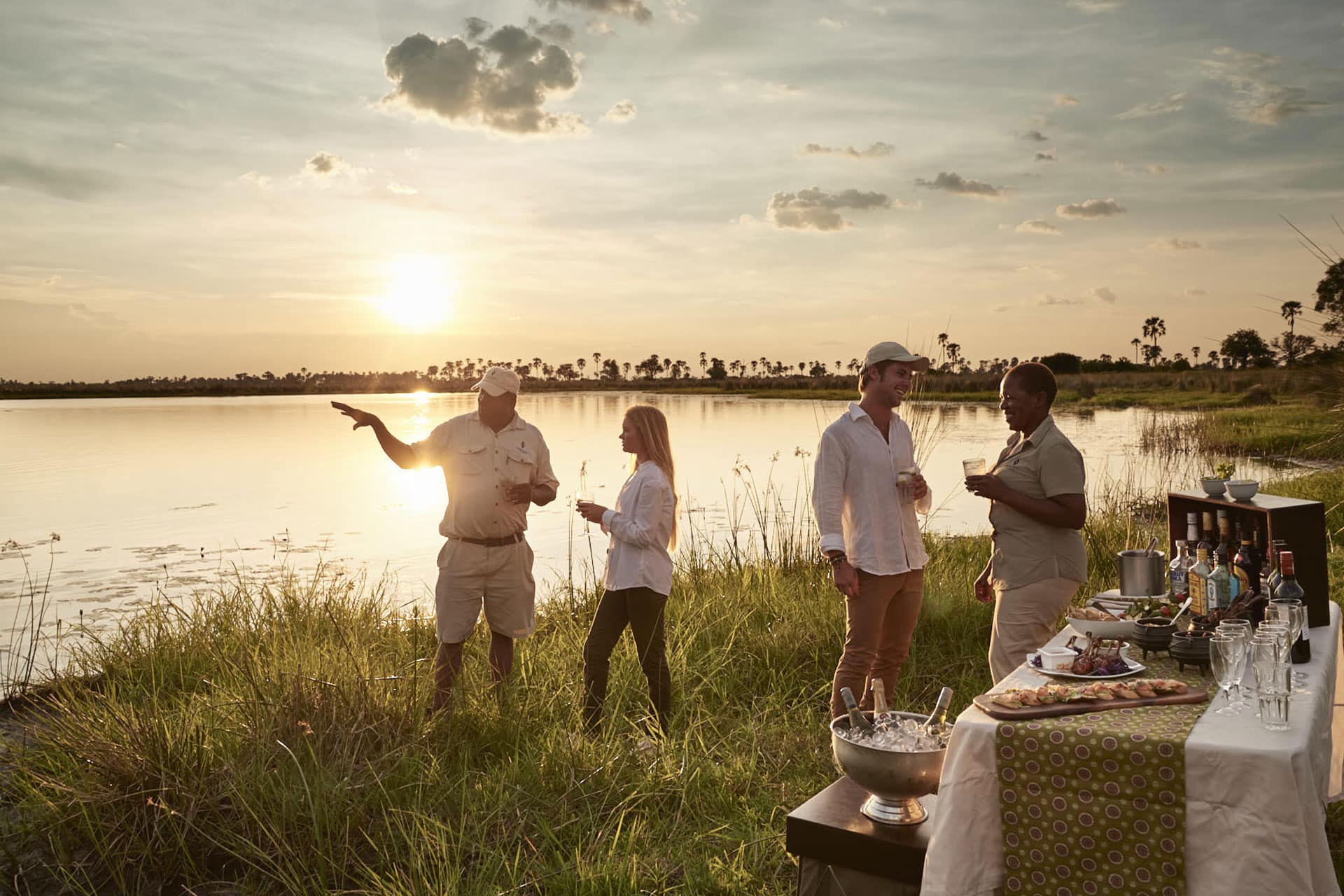People alongside a body of water watching the sun set and having drinks before a night safari at Sanctuary Baines&rsquo; Camp. 