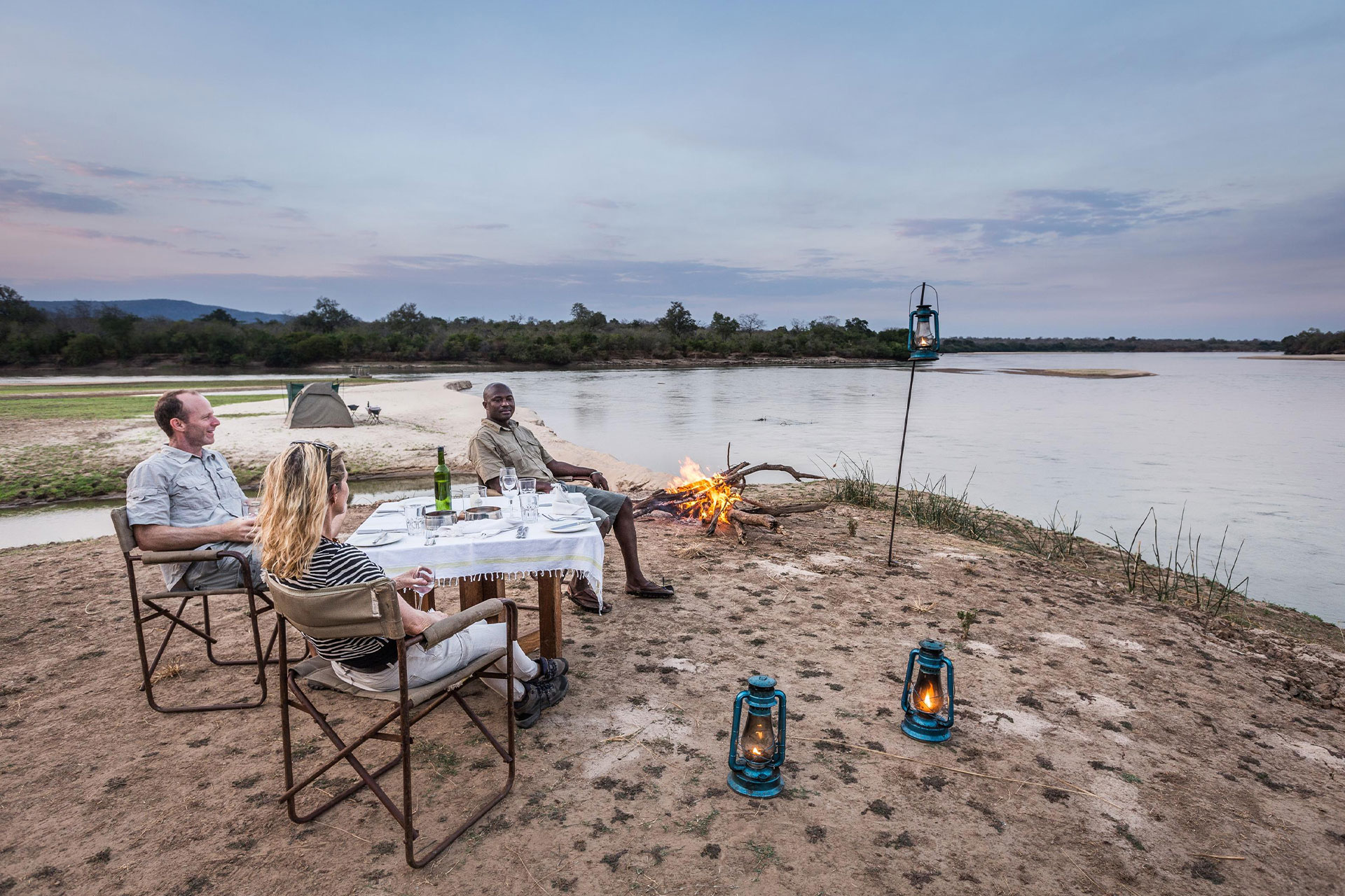 Three people sitting around a table alongside a river with a campfire at Sands River Selous during a night safari.