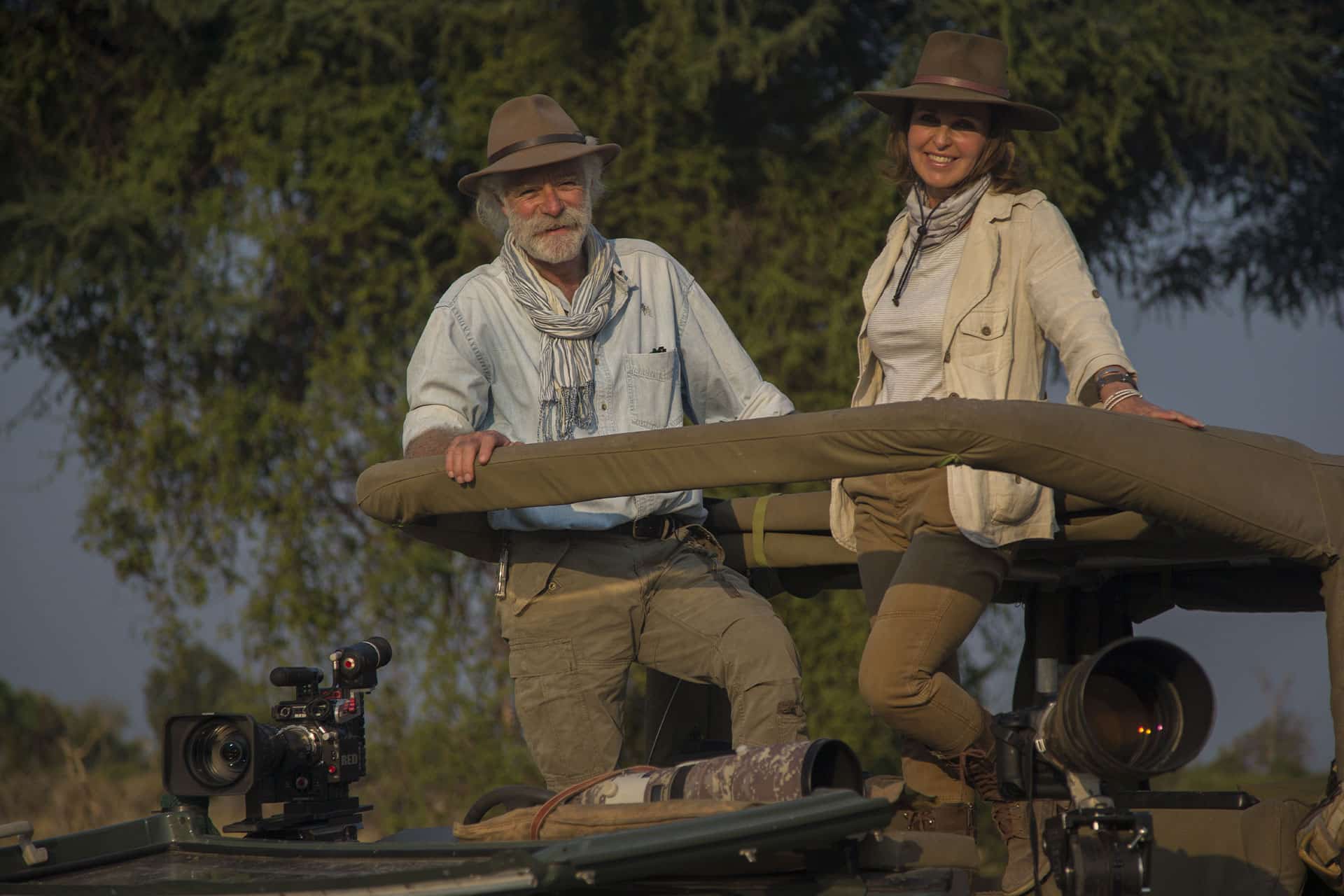 Photographers and filmmakers Dereck and Beverly Joubert standing in an open air safari vehicle smiling. 