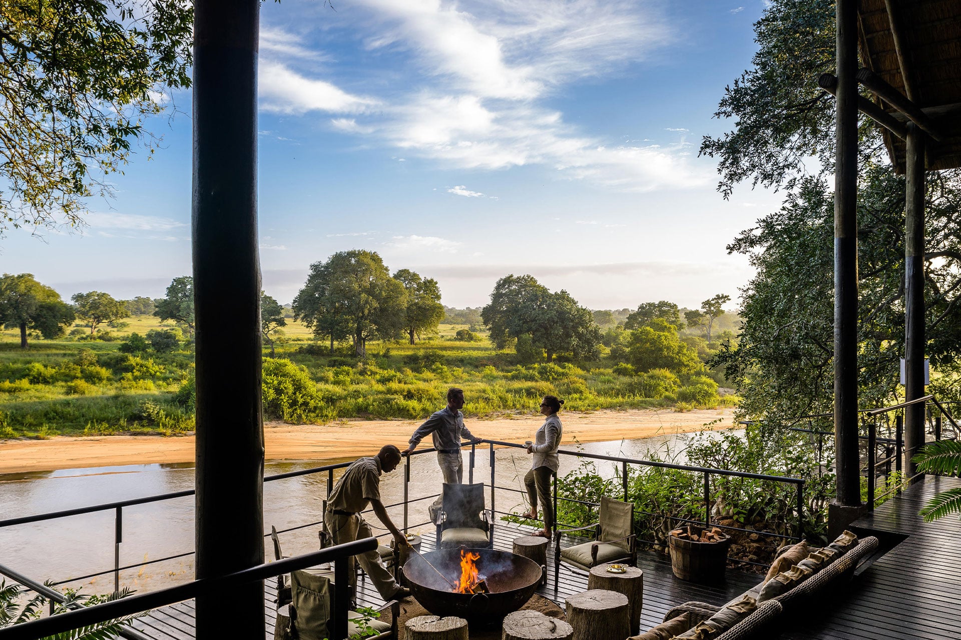 People gathered around the fire pit on the wooden deck with views of the Sand River at Singita Sabi Sands Ebony Lodge. 