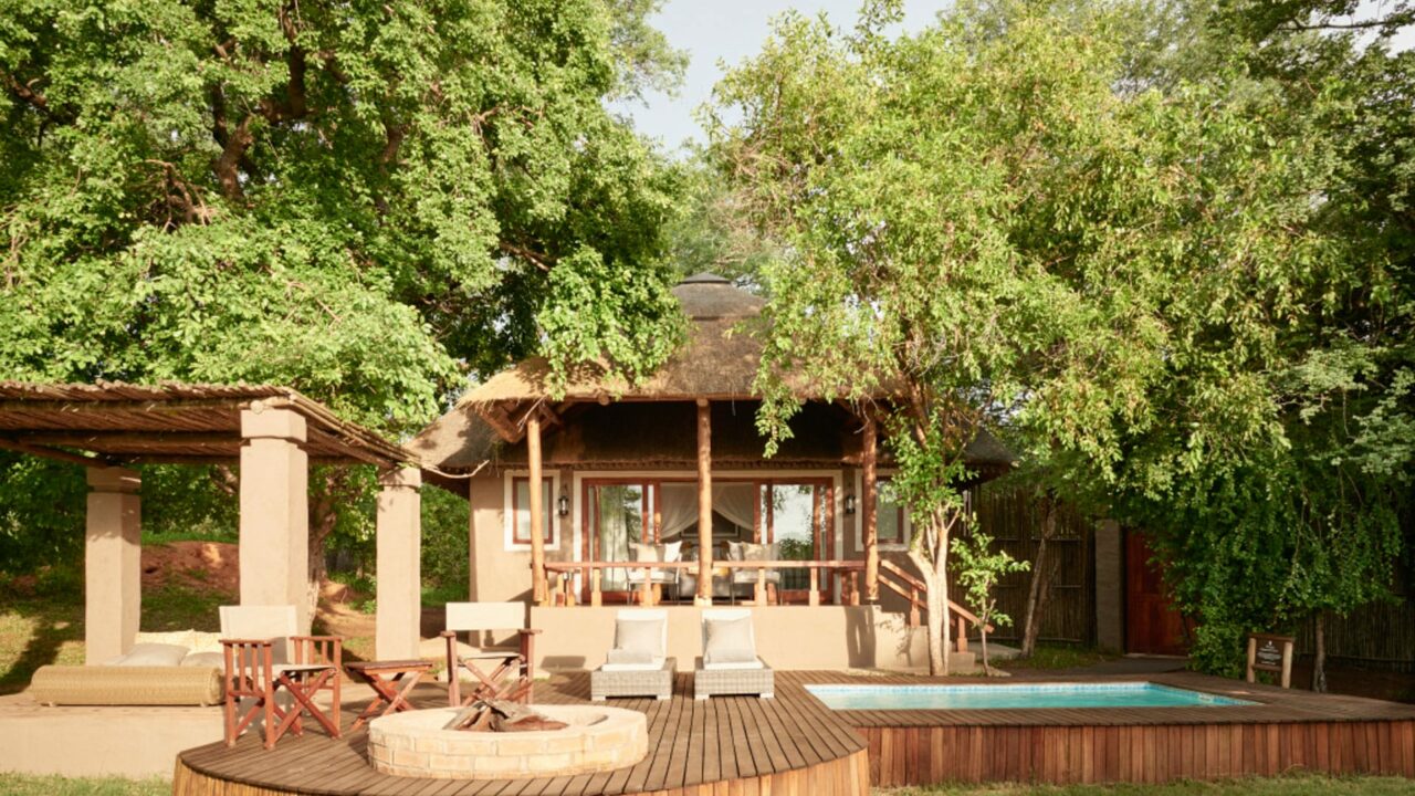 Chobe Chilwero Lodge in Botswana with a thatched-roof building, wooden deck, lounge chairs, fire pit, and small pool surrounded by lush greenery.