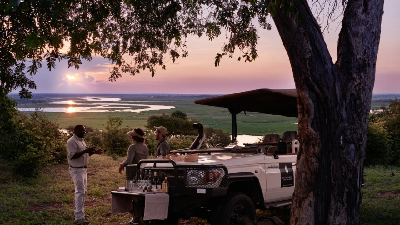 One guide and two tourists standing beside a safari jeep under a tree at sunset, overlooking a scenic river landscape, in the Chobe National Park, in Botswana.