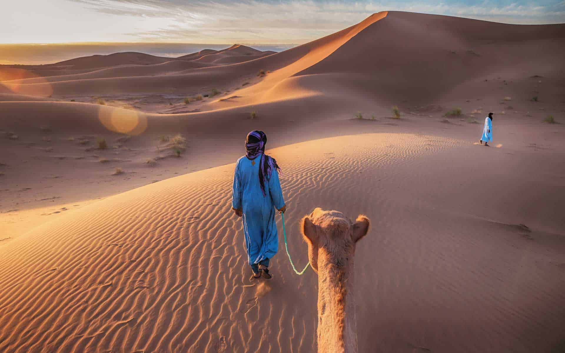 A man leading his camel over the sand dunes of the Sahara Desert - one of the Seven Natural Wonders of Africa